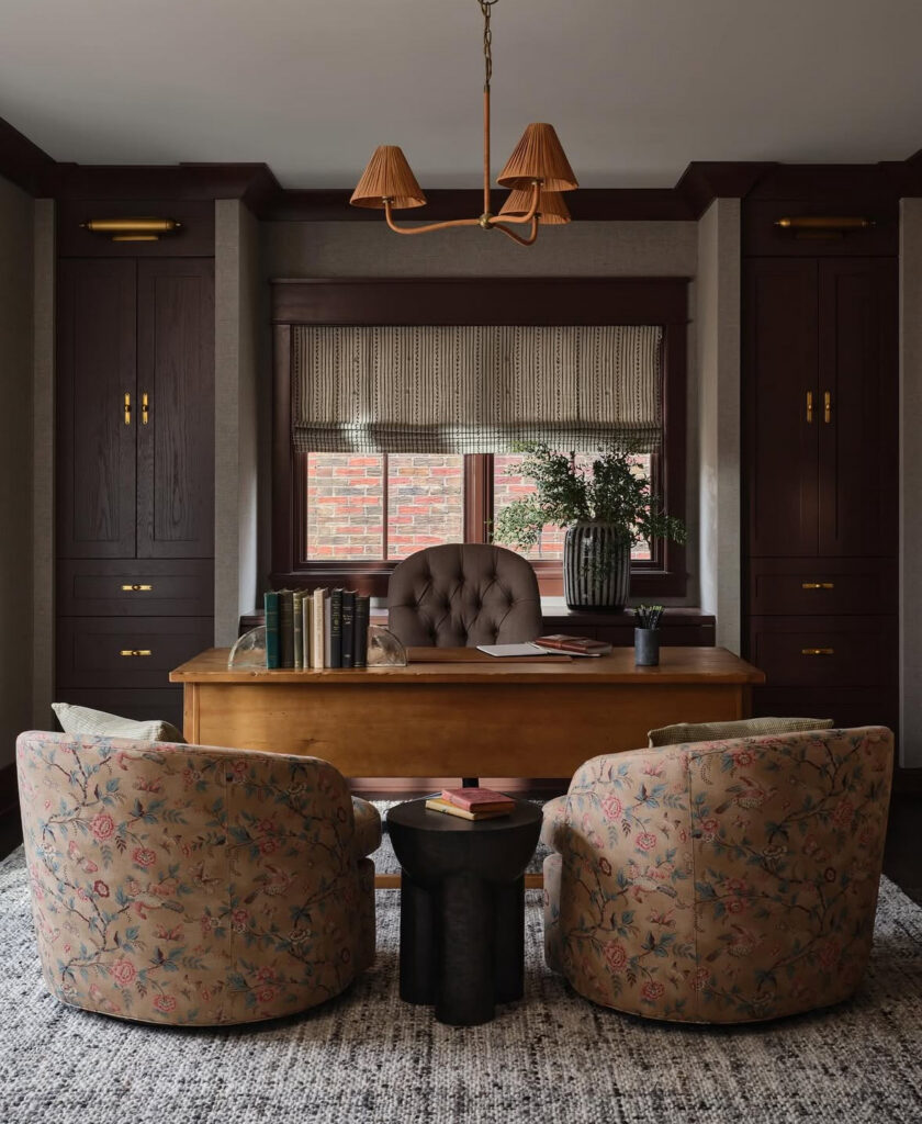 Cozy masculine home office with warm wood desk, tufted desk chair, two floral swivel chairs, burgundy built-ins and a textured area rug