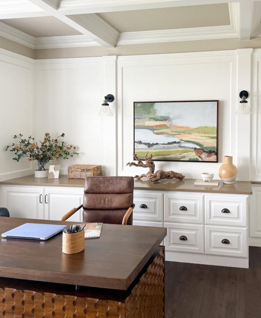 Light-filled masculine home office with white paneling, coffered ceiling, dark wood desk, brown leather chair, long built-in cabinets and colorful landscape artwork