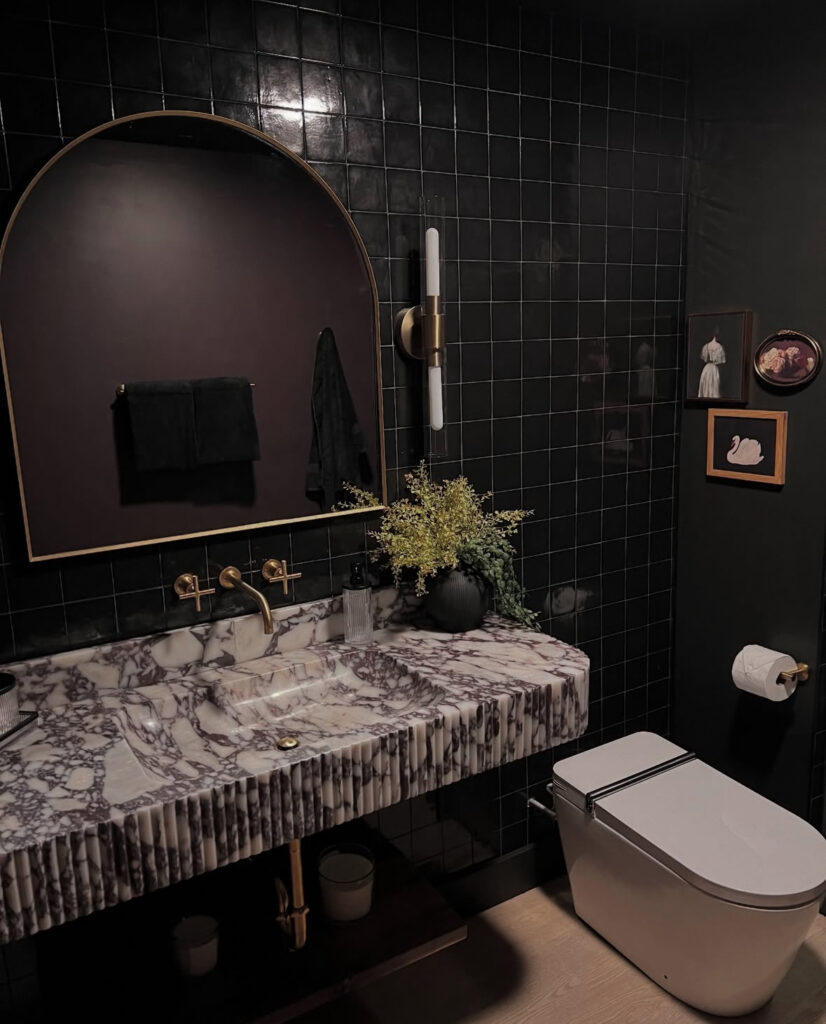 A small bathroom with glossy black tiles, brass accents, and a marble sink showcasing moody bathroom decor.