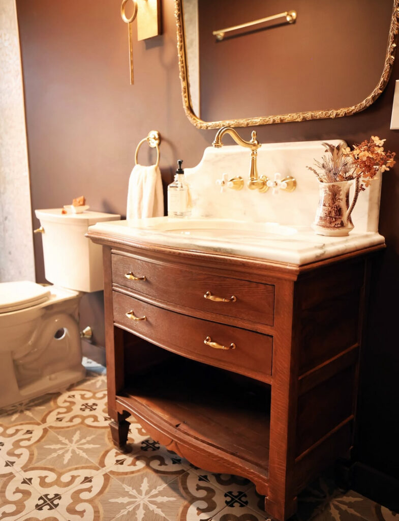 Deep brown walls frame an antique dresser converted into a marble-topped vintage bathroom vanity with brass faucet.
