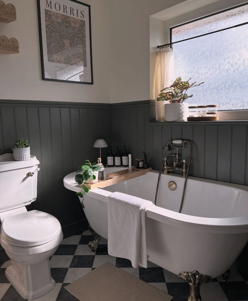 Clawfoot tub, dark beadboard, and framed art create a moody vintage bathroom with plants and bath tray.