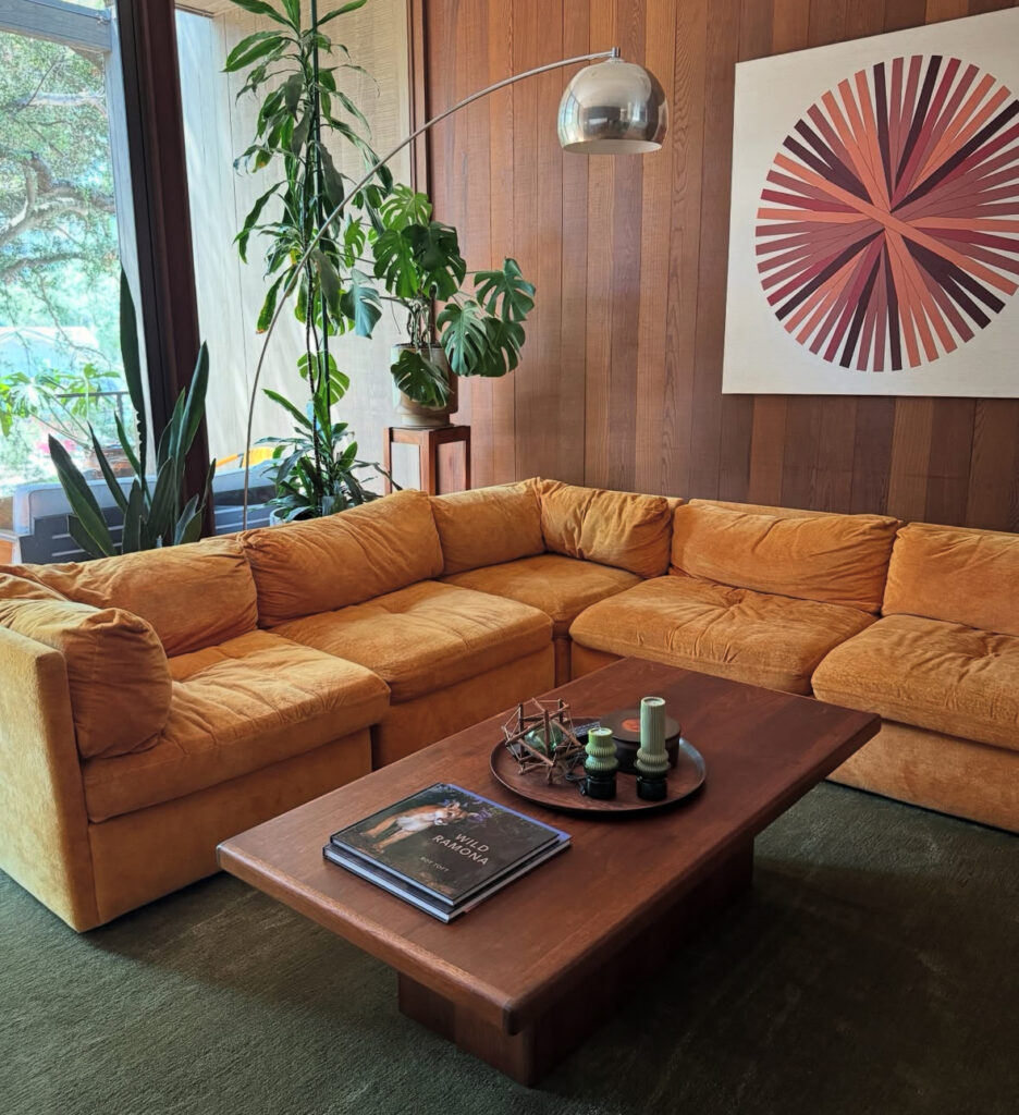 Sunlit 70s inspired living room showcasing a mustard sectional, oversized arc floor lamp, wood paneled walls and large artwork.