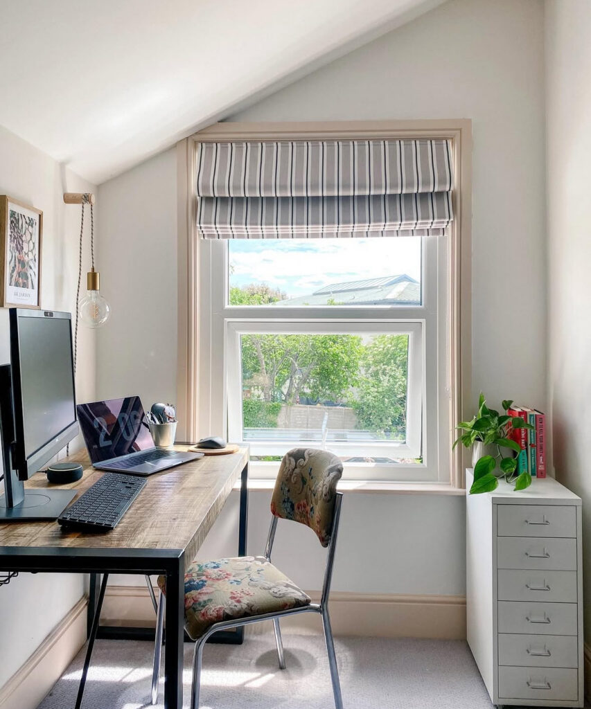 Attic desk by the window with a slim drawer unit, striped shade, and a simple chair, natural light office vibes.