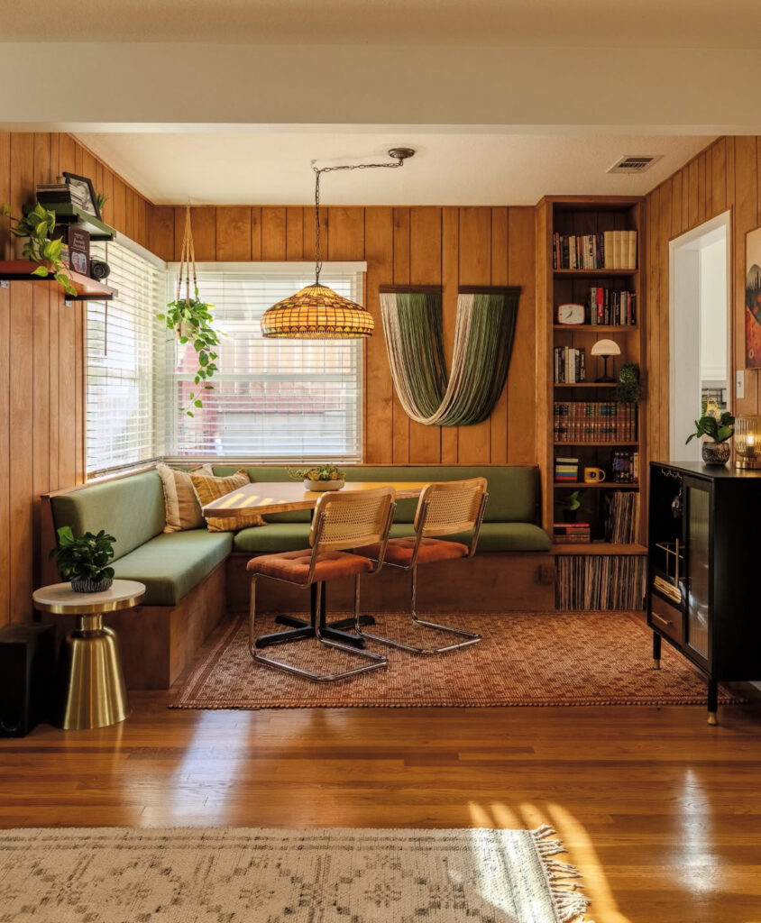 Small dining corner in living room 70s home decor style, with paneled walls, green banquette, cane chairs and library shelves.