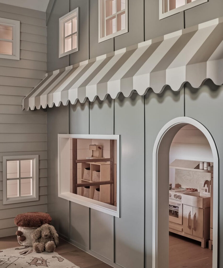 Sage-green pretend play storefront wall with a striped awning, arched doorway, and built-in cubbies creating a whimsical playroom zone.