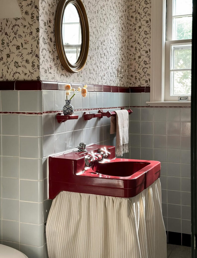 Bold red vintage bathroom sink paired with pale blue wall tile and floral wallpaper.