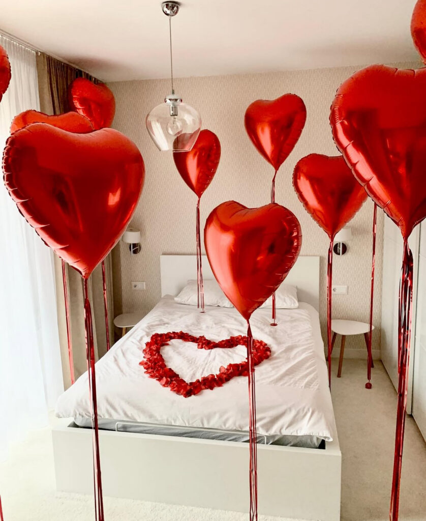 White hotel-style bedroom filled with red heart balloons and rose petals for luxurious valentines day bedroom ideas.