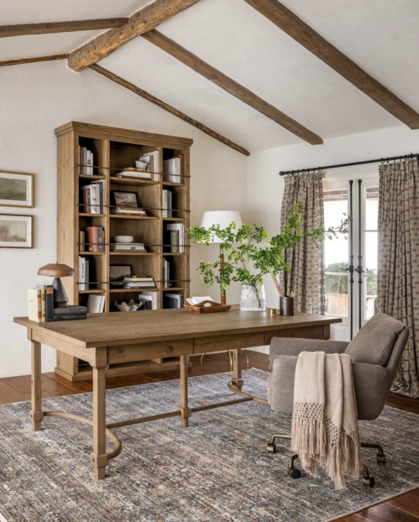 Rustic office decor for women with exposed ceiling beams, tall wood bookcase, large farmhouse style desk, gray upholstered swivel chair and patterned area rug