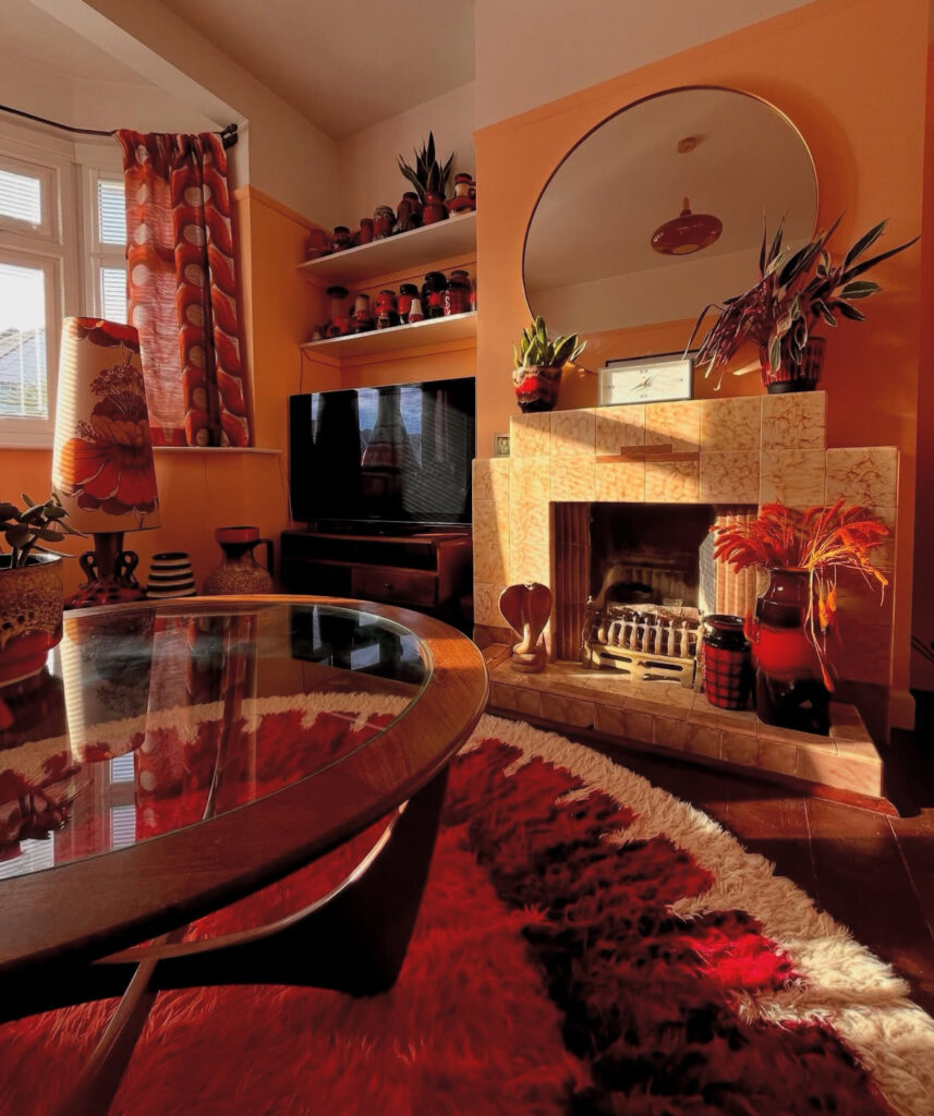 Warm 70s decor living room with orange walls, patterned curtains, glass coffee table and fluffy red and white shag rug by the fireplace.