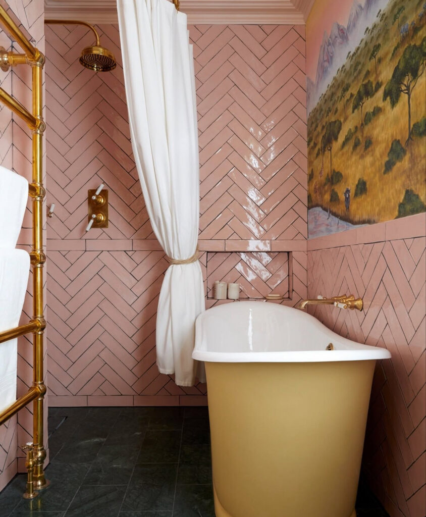 Vintage bathroom with soft pink herringbone tile, pastel colors, brass fittings, and a bold yellow tub with mural detail.