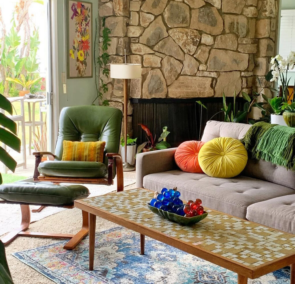 Playful 70s decor living room with stone fireplace, green leather lounge chair, gray sofa and round colorful cushions.