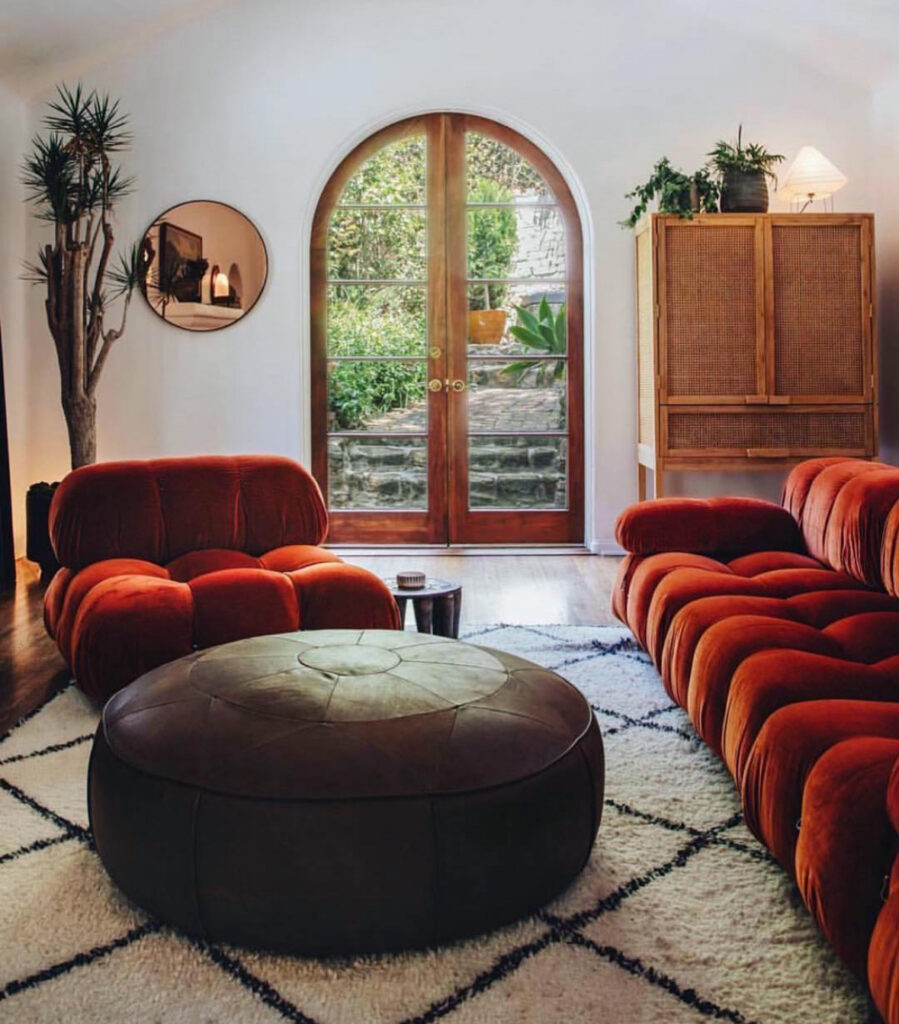 Chic 70s inspired living room with rust tufted modular sofas, oversized round ottoman and arched French doors to the garden.