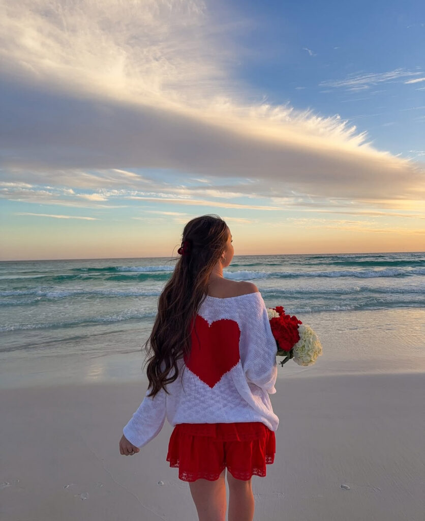 Valentine’s Day outfit at the beach: off-the-shoulder white heart sweater with a large red heart on the back, red shorts, and a bouquet of roses at sunset.
