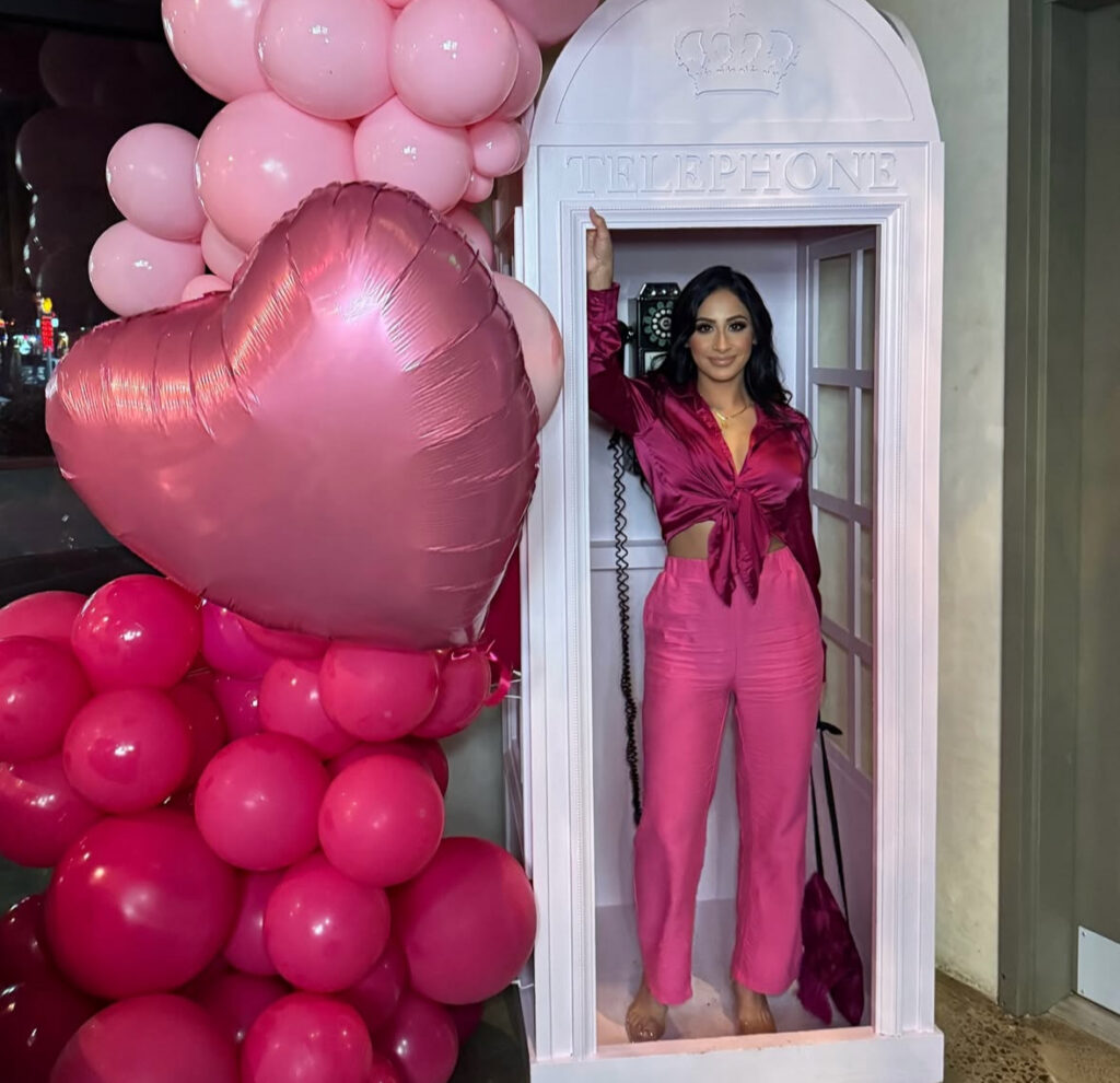 Extra-in-the-best-way Valentines Day outfit: monochrome pink look in a telephone booth with a heart balloon display.