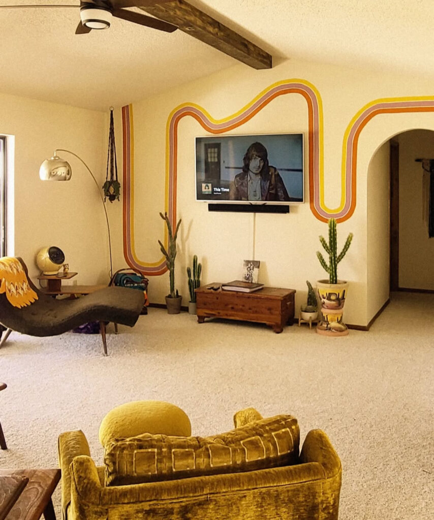 Relaxed living room 70s home decor with painted rainbow stripe behind the TV, mustard chair, cacti and vintage chaise.