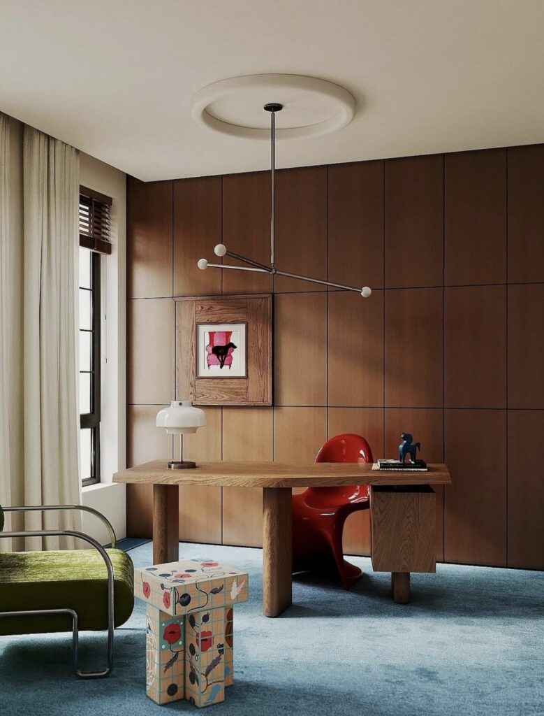 Warm wood paneled home office with blue carpet, chunky oak desk, red modern chair, green velvet armchair and a small patterned pedestal table.