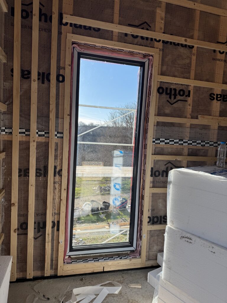 Tall narrow window installed inside timber frame wall with pink airtight window tape around the reveal