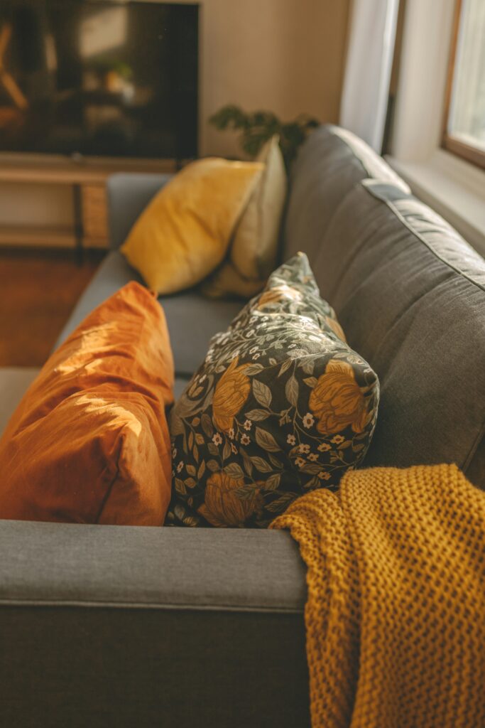 Close-up of a gray sofa styled with a chunky mustard-yellow knit throw blanket, a dark floral pillow with golden-orange blooms, and coordinating rust and gold accent pillows.