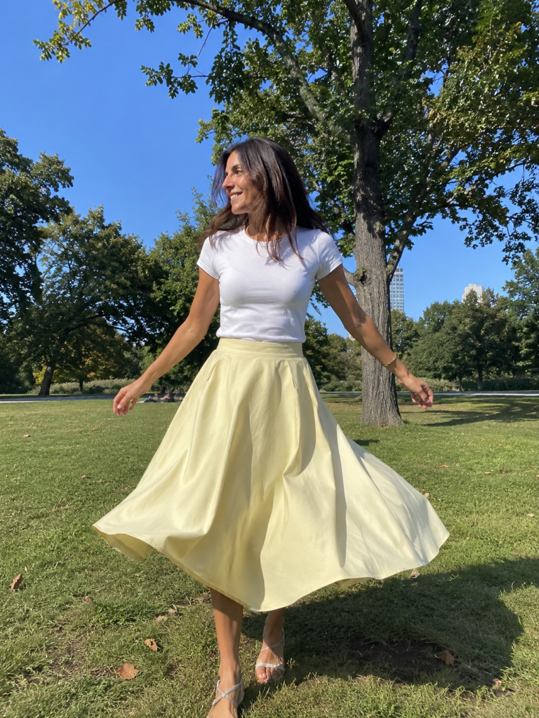 Woman in a butter yellow A-line skirt and white tee twirling in a sunny park, summer work outfit