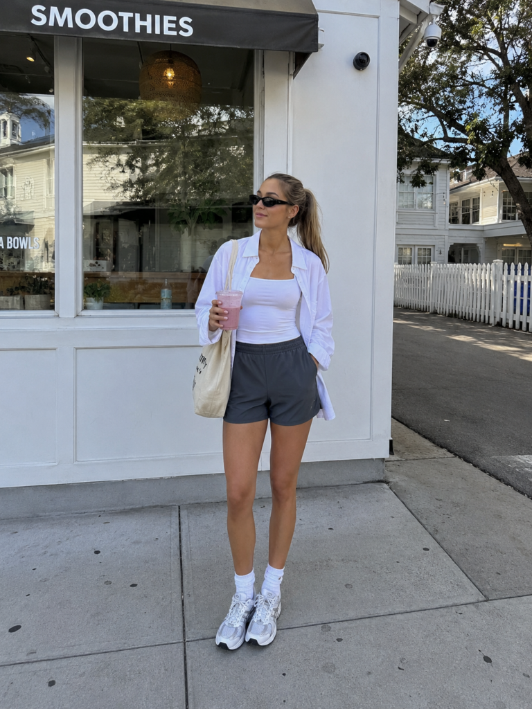 Sporty casual outfit with gray athletic shorts, a white tank top, oversized white shirt, and sneakers outside a smoothie shop.