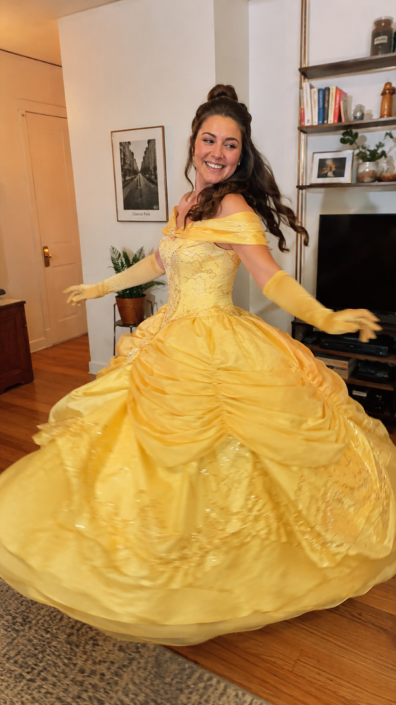 Woman in a flowing yellow ballgown twirling with arms out, Belle Disney Halloween costume