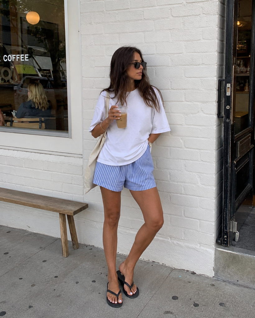 Casual summer outfit with striped boxer shorts, an oversized white t-shirt, flat sandals, and a canvas tote bag.