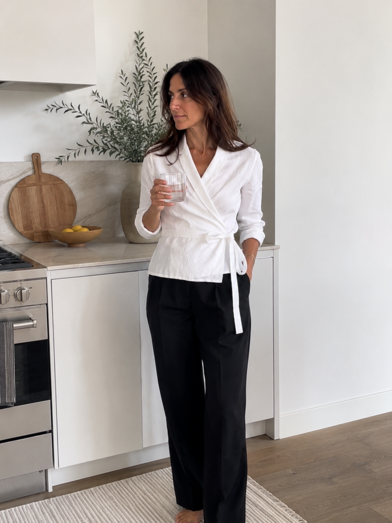 Woman in a white linen wrap top and black trousers in a bright white kitchen, summer work outfit