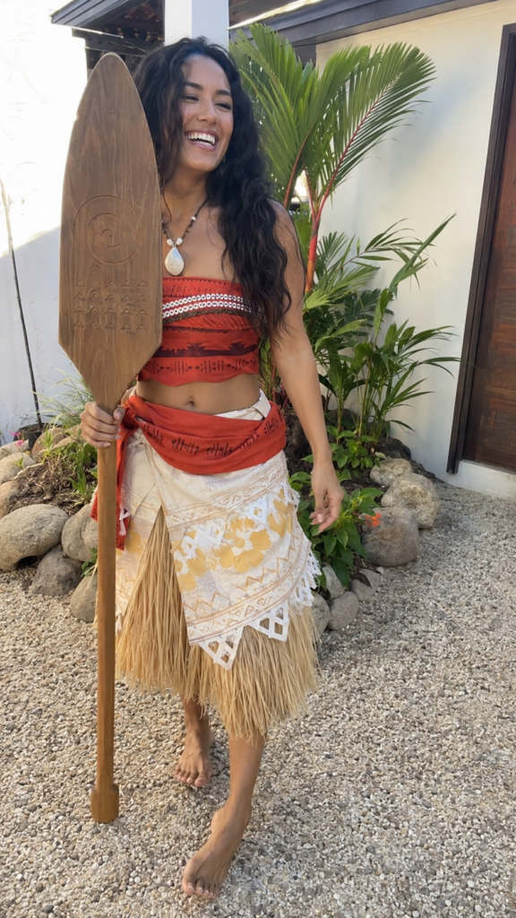 Woman in a tropical two-piece with a grass skirt holding a wooden oar, Moana Disney Halloween costume