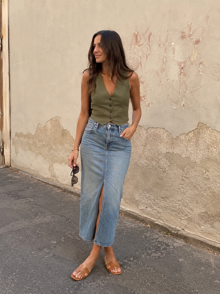Woman wearing an olive green vest top with a denim midi skirt and brown leather sandals near a textured wall.