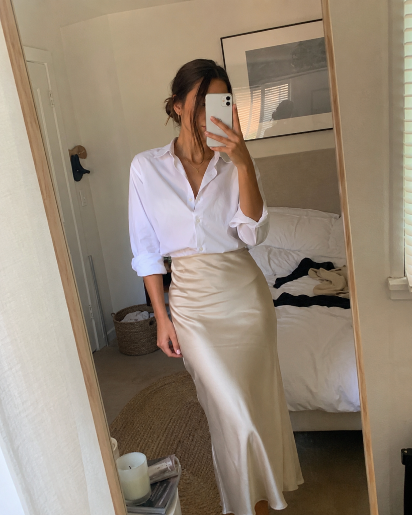 Woman in a champagne satin slip skirt and white button-down taking a bedroom mirror selfie, summer work outfit