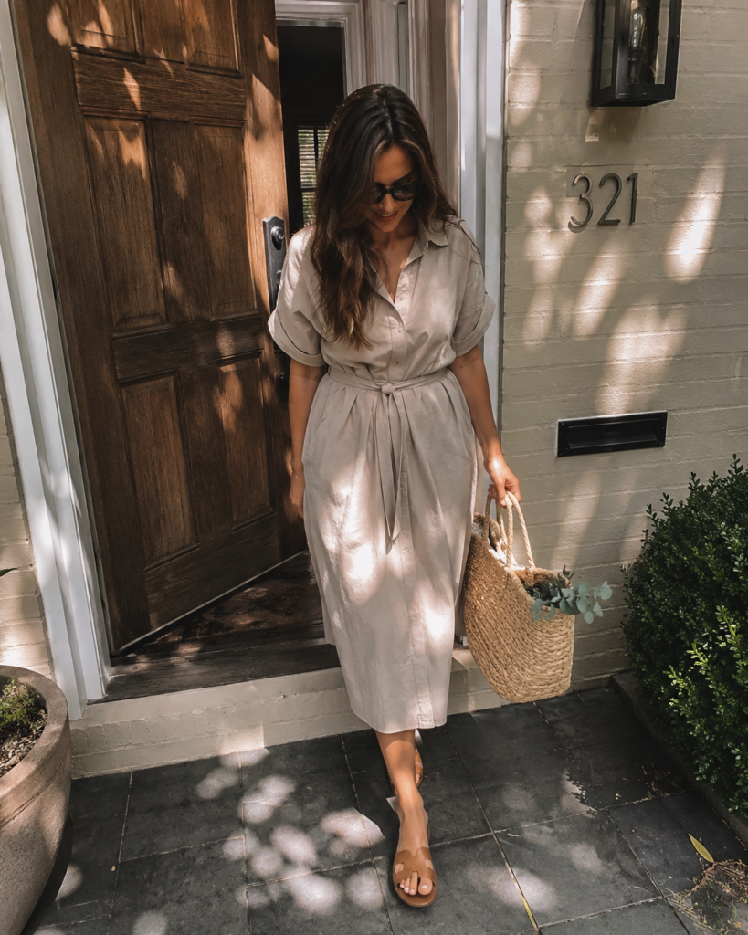 Woman in a belted beige cotton shirtdress and tan sandals walking out onto a porch, summer work outfit