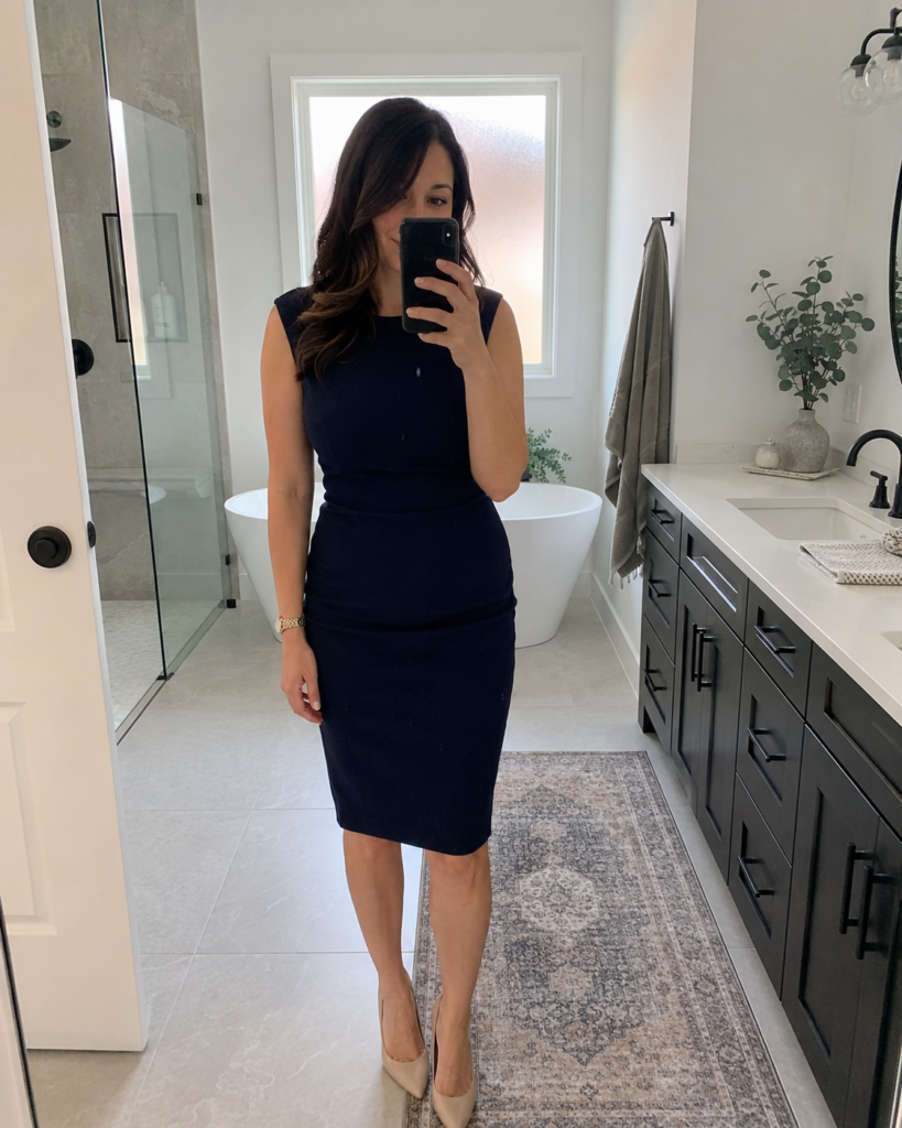 Woman in a sleeveless navy sheath dress and nude pumps taking a bathroom mirror selfie, summer work outfit