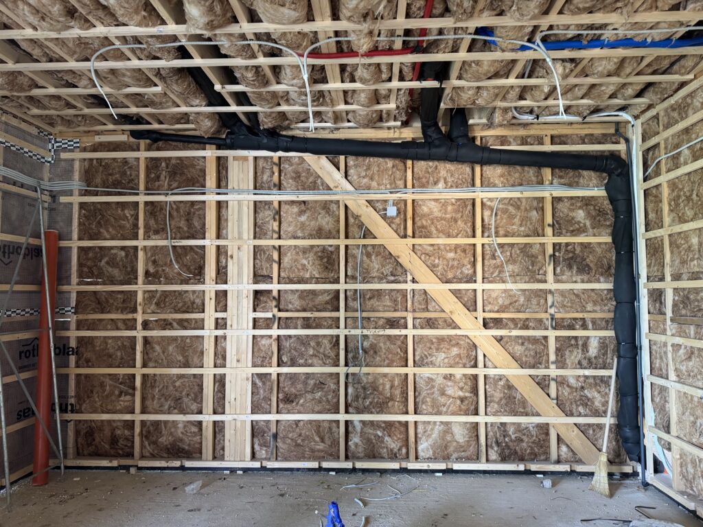 Insulated stud wall with electrical cables run to both sides of the room for the TV, black drain pipe crossing the ceiling above