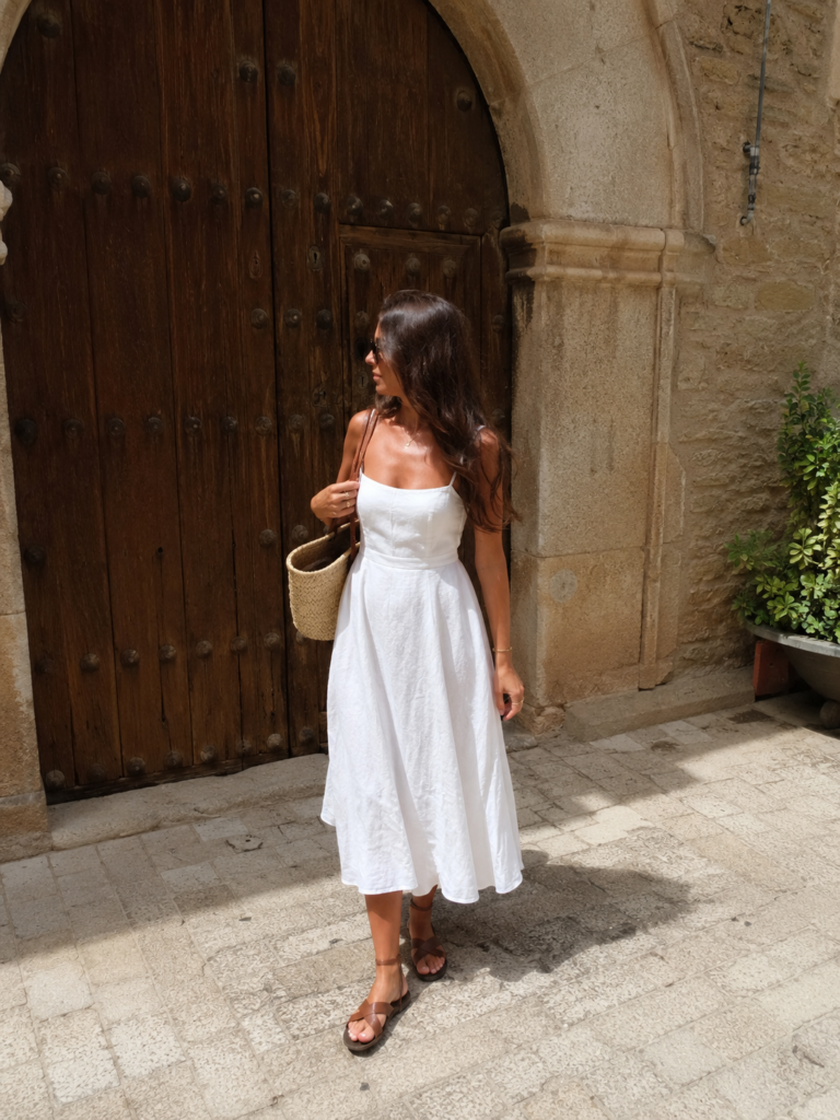 White cotton dress styled with brown leather sandals and a woven bag near a rustic doorway.