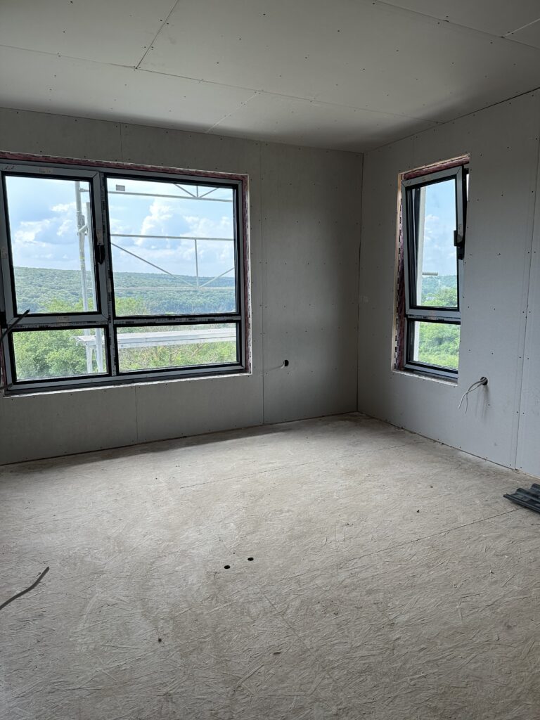 Second-floor bedroom with grey drywall up, two black-framed windows overlooking green hills, and a bare OSB subfloor