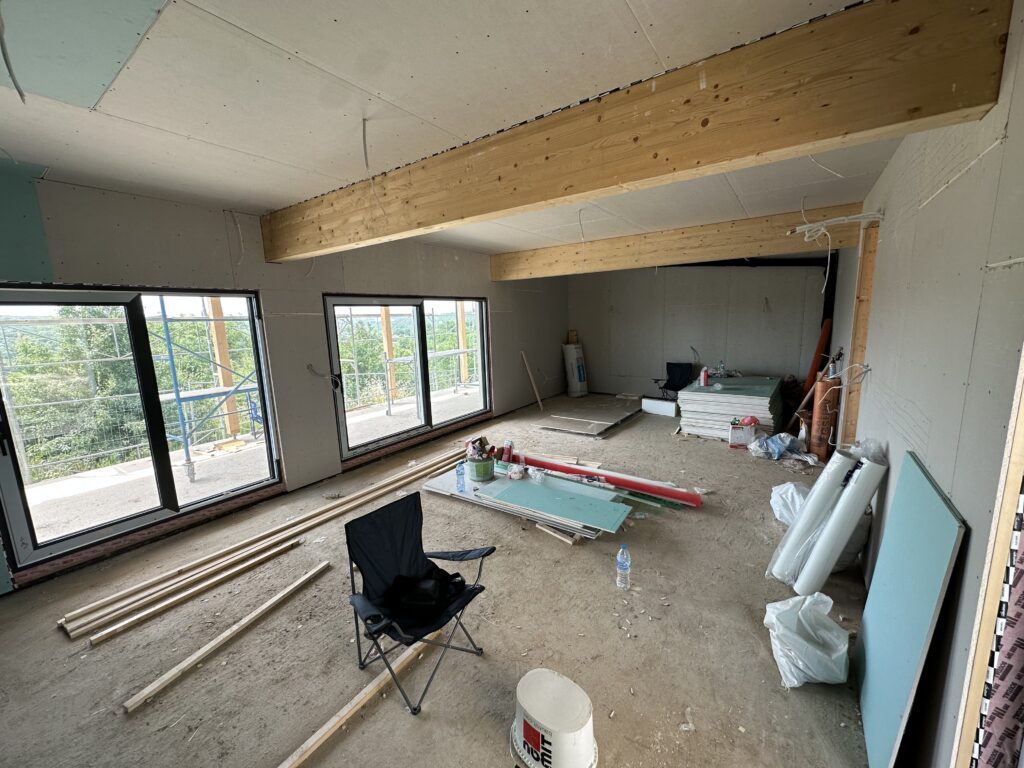 Open-plan living room with drywall going up, an exposed timber beam across the ceiling, big black windows, and building materials on the floor