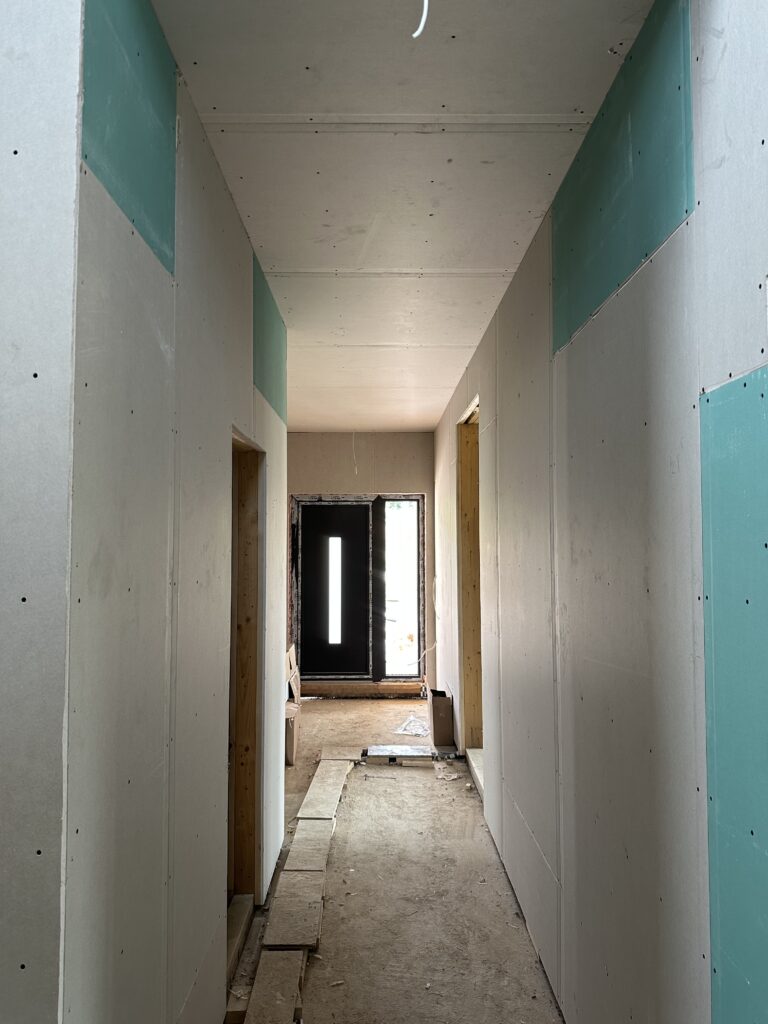 Drywalled corridor with green and grey board, looking toward the black front door with its vertical glass slot and the stairwell beyond