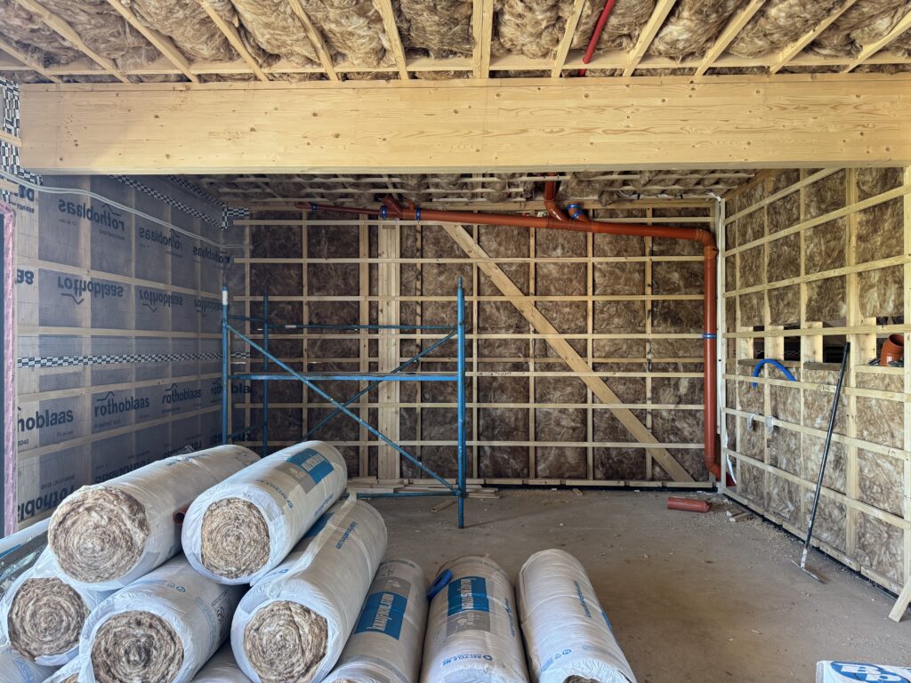 Room mid-build with mineral wool insulation in the walls and ceiling, an orange drain pipe overhead, scaffolding, and rolls of insulation stacked on the floor
