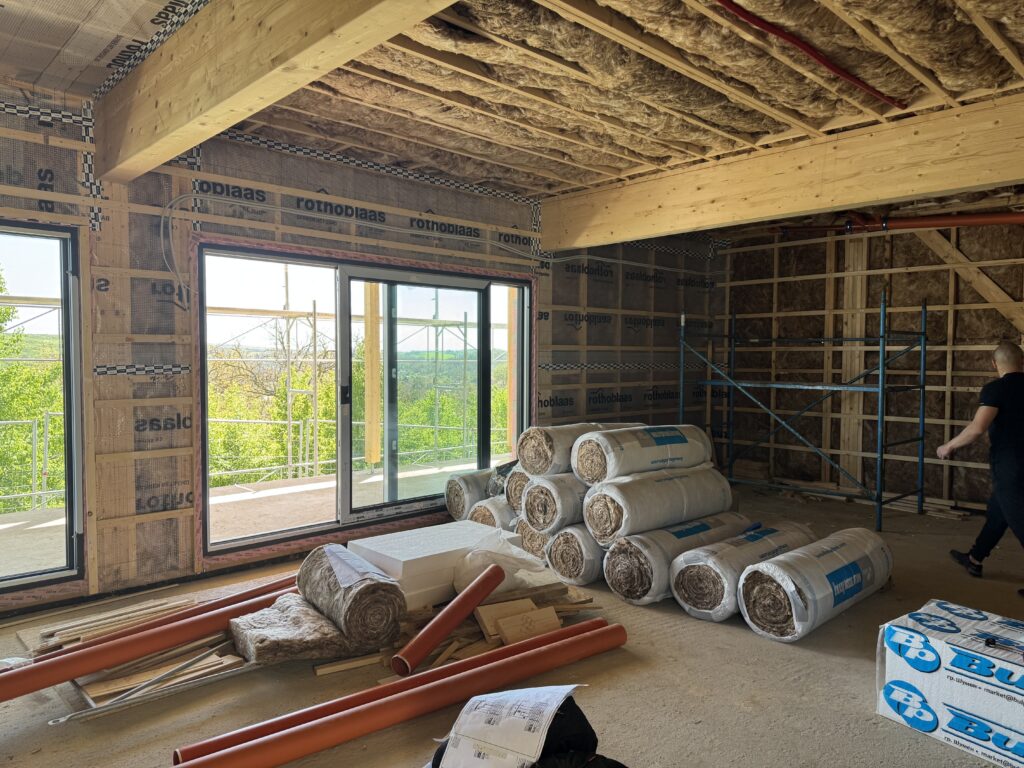 Living room under construction with a large glulam beam, insulated walls and ceiling, big black windows over the countryside, and insulation rolls and orange pipe on the floor
