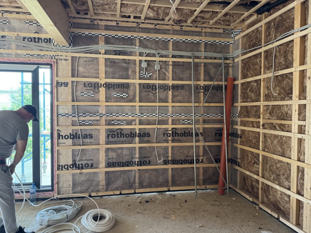 Timber-frame living room wall with electrical cables and outlet boxes roughed in over the Rothoblaas membrane, a worker pulling cable on the left, and coils of cable on the floor