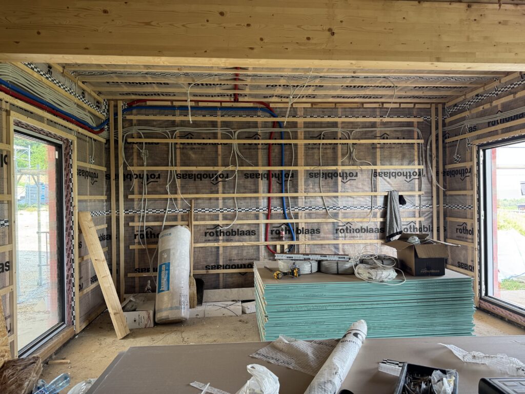 Living room timber-frame walls with red and blue water lines and white electrical cables roughed in across the membrane, drywall sheets stacked ready to install