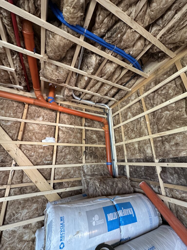 Corner of insulated timber framing with an orange drain pipe and blue and red water lines routed through the studs and ceiling