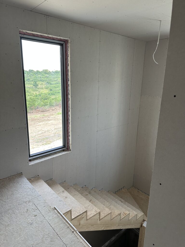 Drywalled stairwell with a tall narrow window and cut timber stair stringers waiting for treads