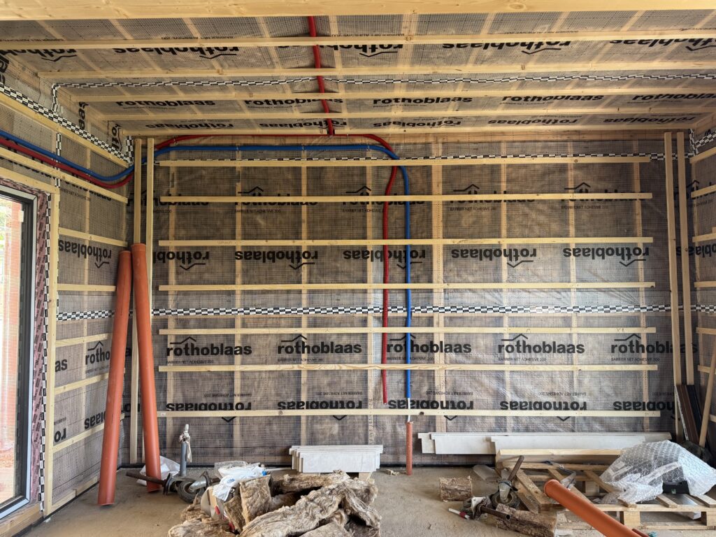 Timber-frame wall with red and blue hot and cold water lines running across the Rothoblaas membrane, orange drain pipes leaning in the corner