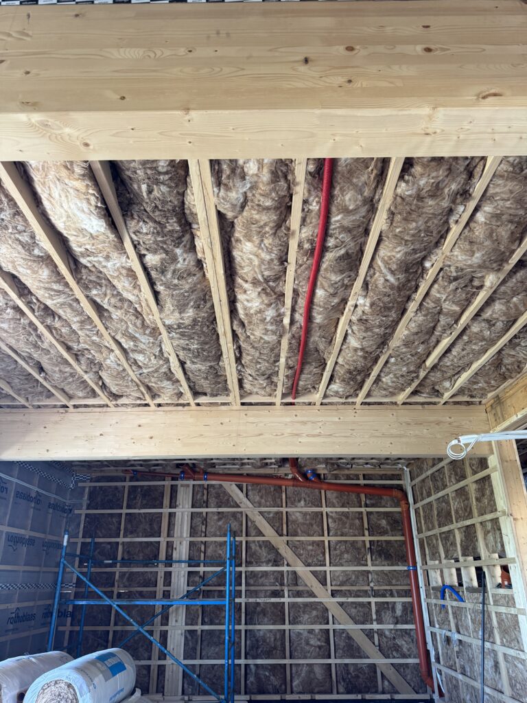 Ceiling joists packed with mineral wool insulation with a red hot water line running through, part of the home's hot water recirculation system