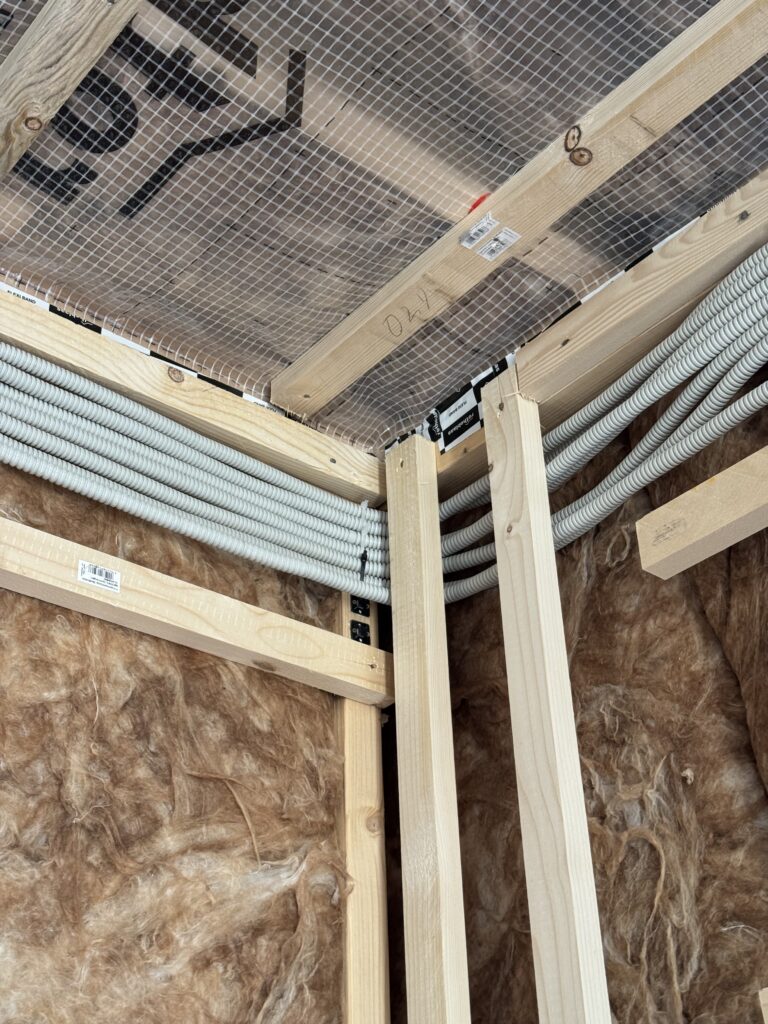 Close-up of bundled white electrical conduit and cables running along the top of insulated timber-frame walls toward the distribution point