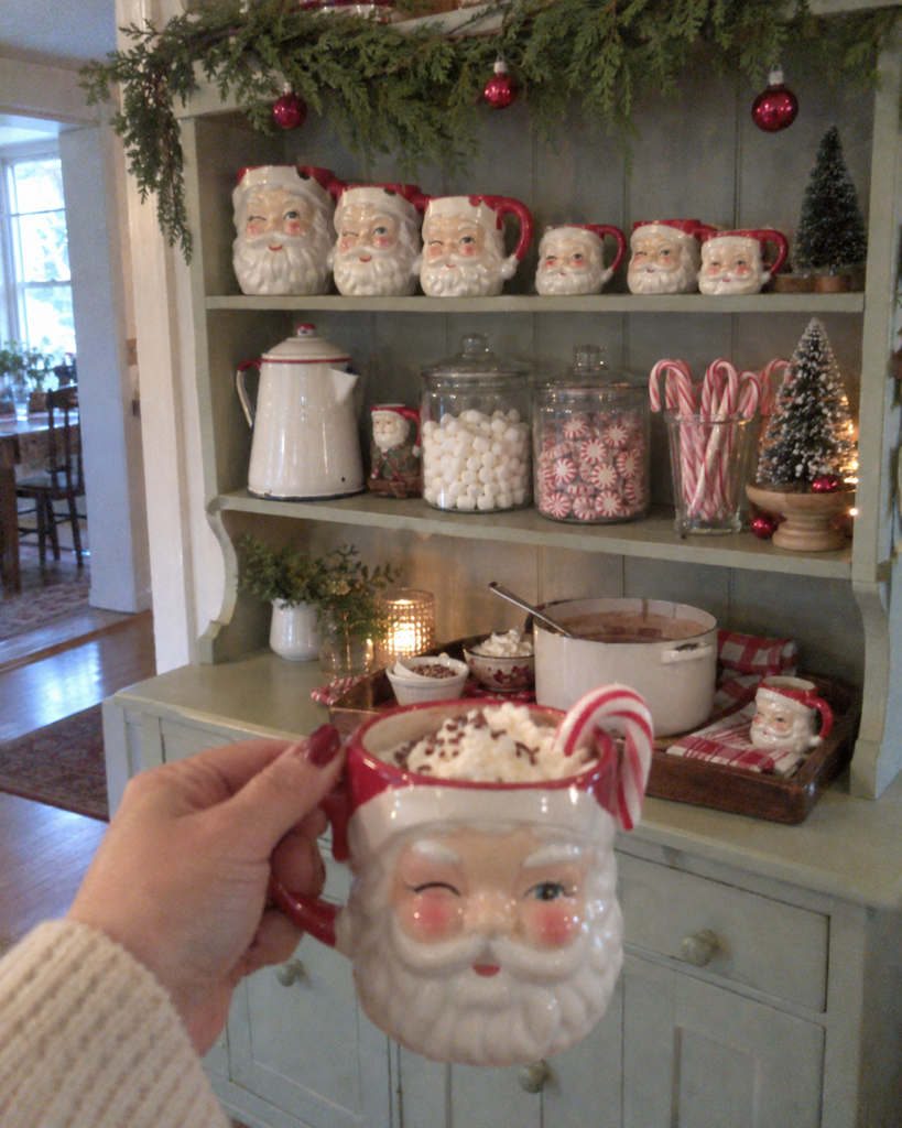 Collection of vintage ceramic Santa mugs displayed on open kitchen shelves beside a festive hot cocoa station
